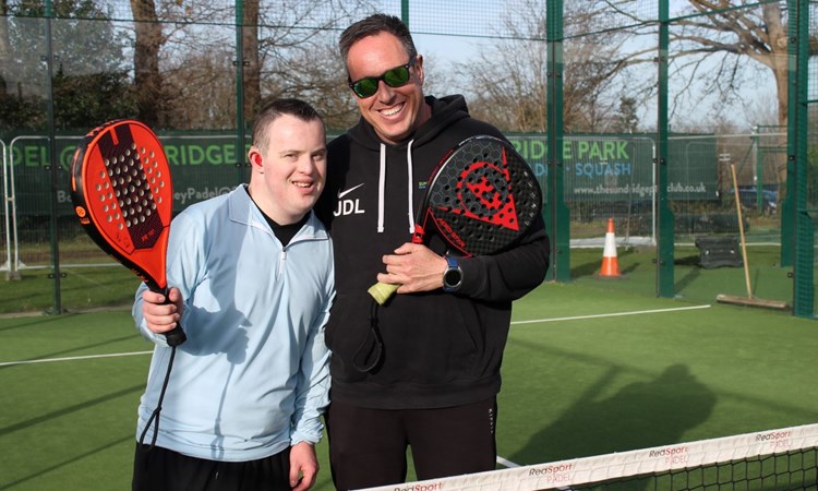 A young man with Downs Syndrome poses with a padel coach on an outdoor padel court