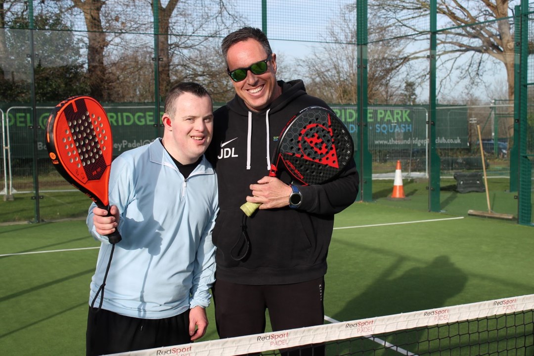 A young man with Downs Syndrome poses with a padel coach on an outdoor padel court