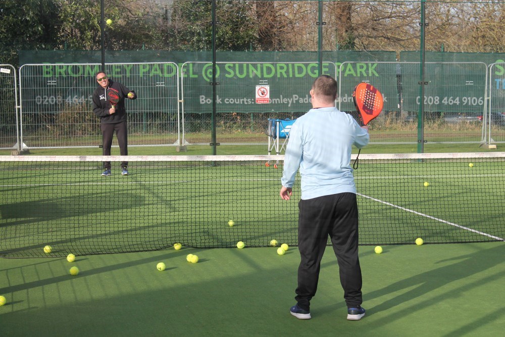 A young man with Downs Syndrome plays padel with a padel coach on an outdoor padel court