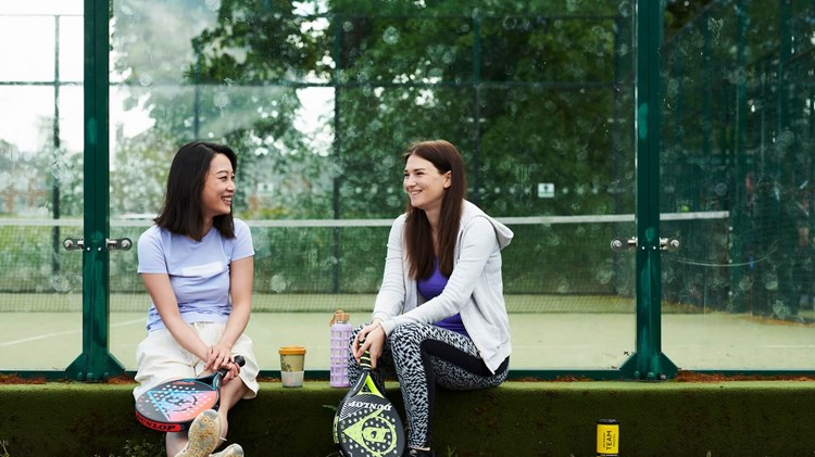 Two friends sitting on the edge of a padel court chatting with their padel equipment round them