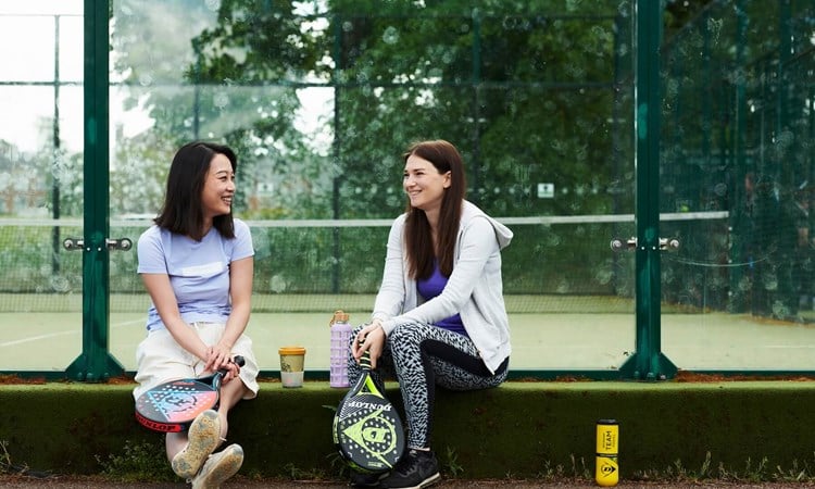 Two friends sitting on the edge of a padel court chatting with their padel equipment round them