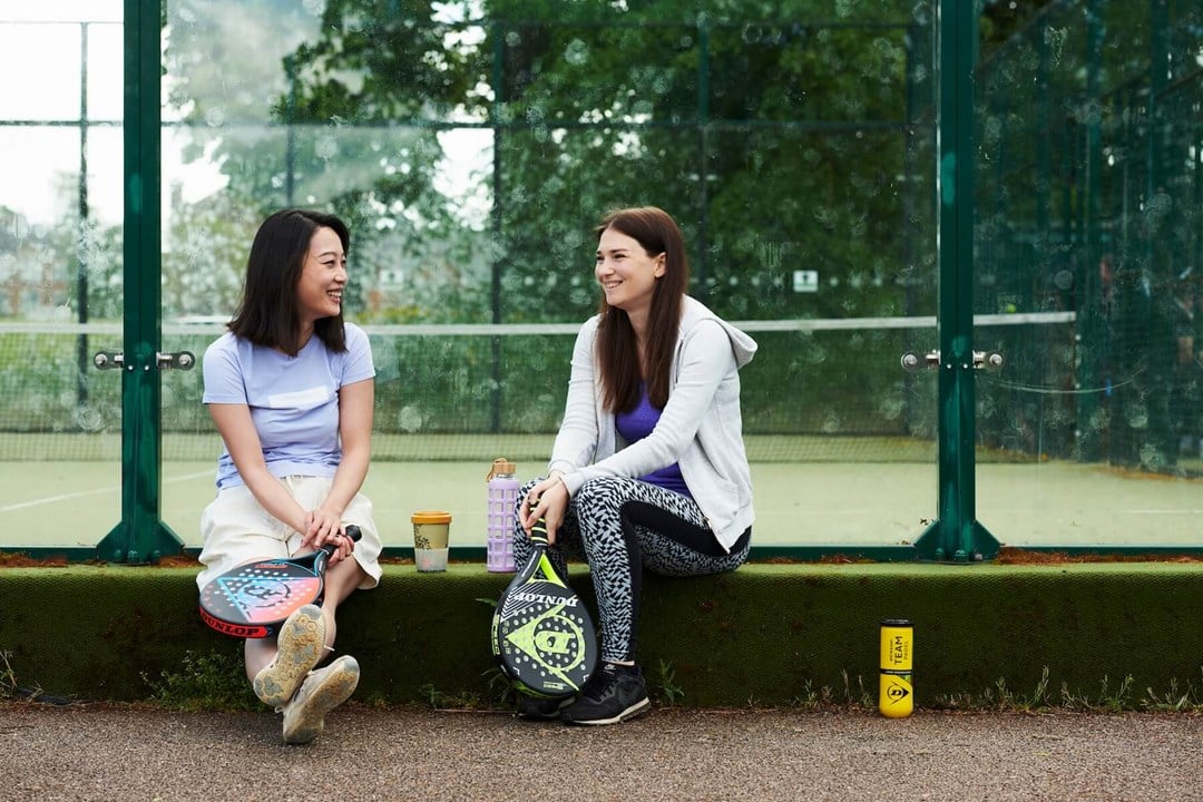 Two friends sitting on the edge of a padel court chatting with their padel equipment round them