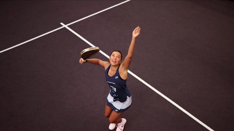An ariel view shot of Catherine rose throwing a ball in the air and preparing to hit it with her padel racket on a padel court