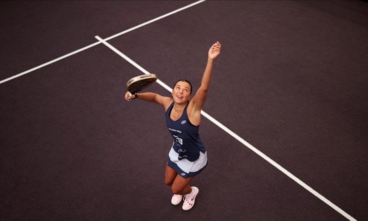 An ariel view shot of Catherine rose throwing a ball in the air and preparing to hit it with her padel racket on a padel court