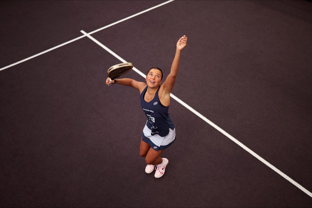 An ariel view shot of Catherine rose throwing a ball in the air and preparing to hit it with her padel racket on a padel court