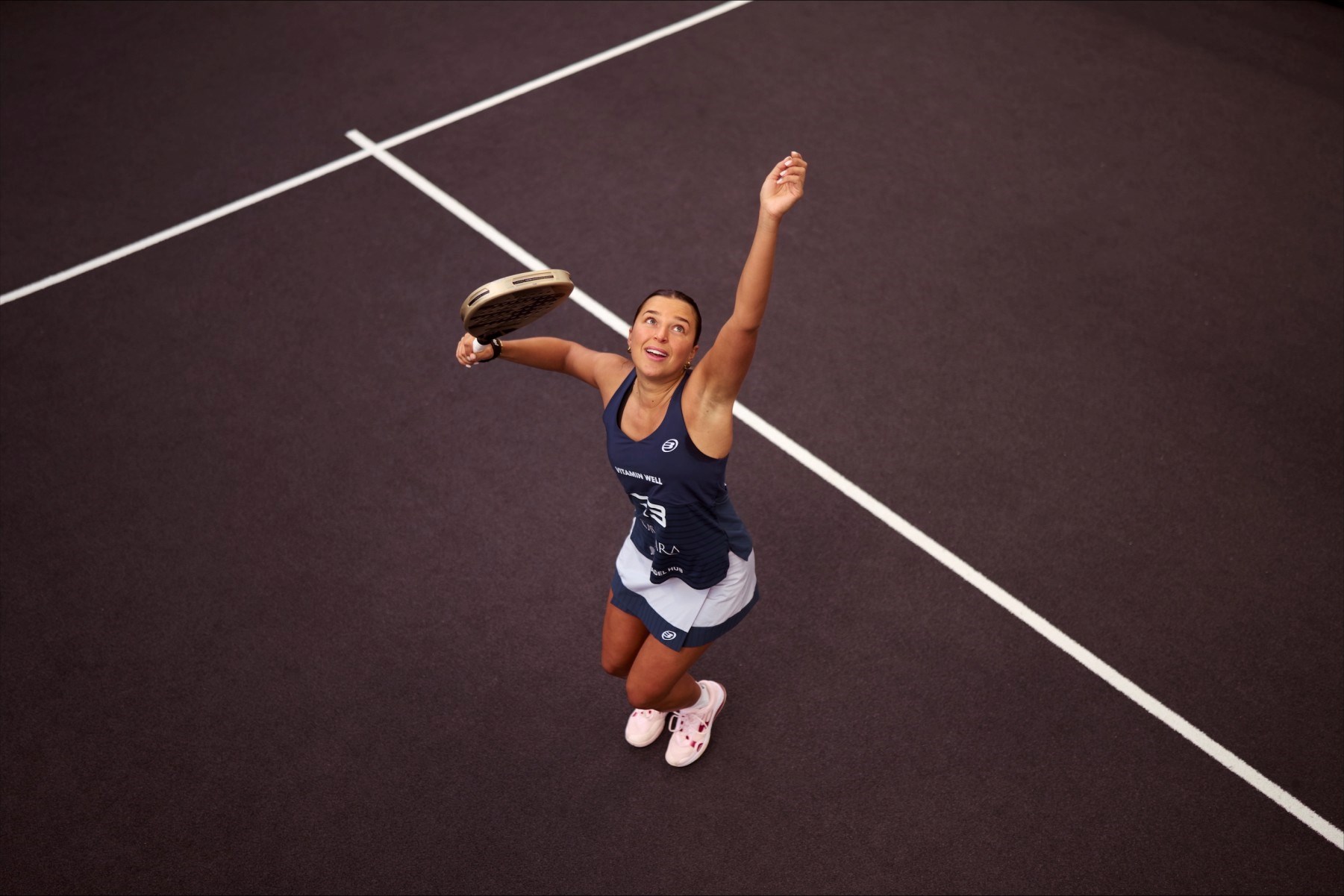 An ariel view shot of Catherine rose throwing a ball in the air and preparing to hit it with her padel racket on a padel court
