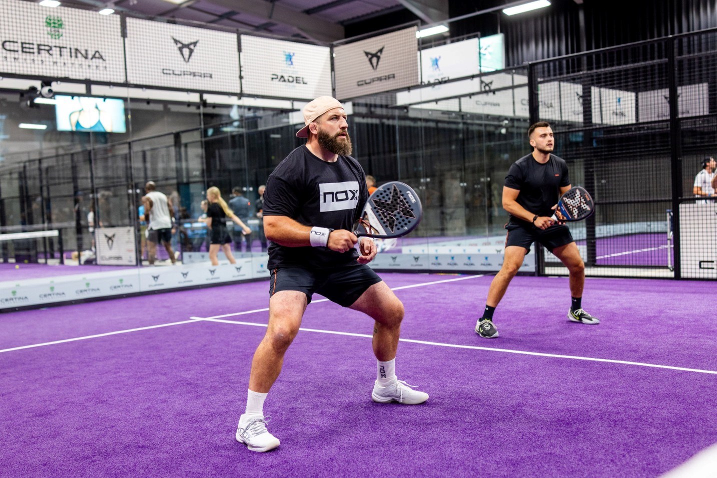 Former England rugby player Joe Marler stood holding a padel bat on a padel court alongside professional padel player Louie Harris