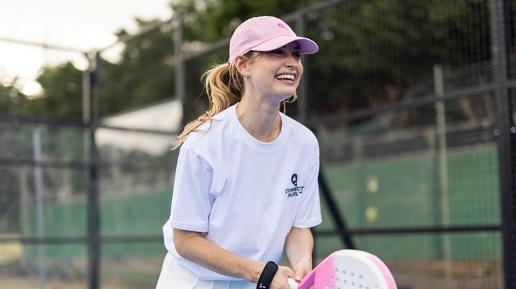 Actress Lily James wearing a white t-shirt saying 'Connect Padel' and a pink hat, holding a pink padel racket and preparing to return a ball on a padel court