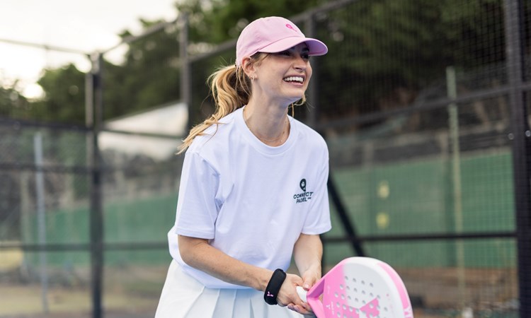 Actress Lily James wearing a white t-shirt saying 'Connect Padel' and a pink hat, holding a pink padel racket and preparing to return a ball on a padel court