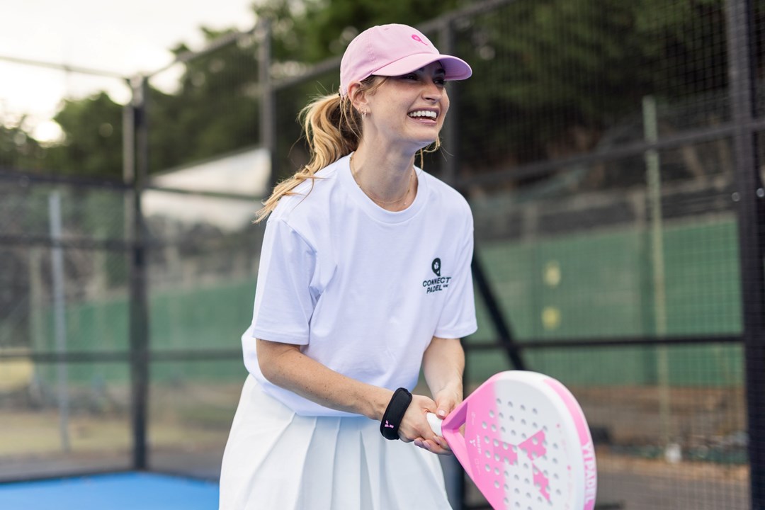 Actress Lily James wearing a white t-shirt saying 'Connect Padel' and a pink hat, holding a pink padel racket and preparing to return a ball on a padel court