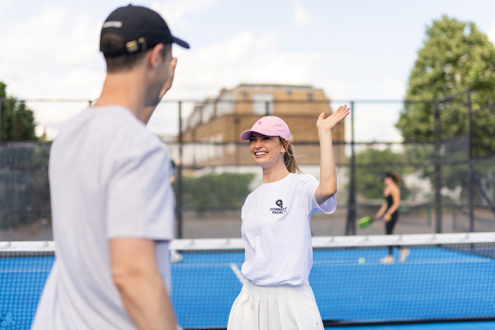 Actress Lily James, wearing a white t-shirt saying 'Connect Padel' and a pink hat, giving a high-five to her brother Charlie on a padel court