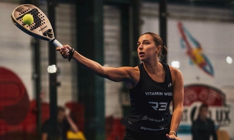 A woman wearing a bacl sports outfit hits a tennis ball with a padel racket