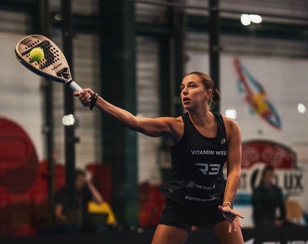 A woman wearing a bacl sports outfit hits a tennis ball with a padel racket