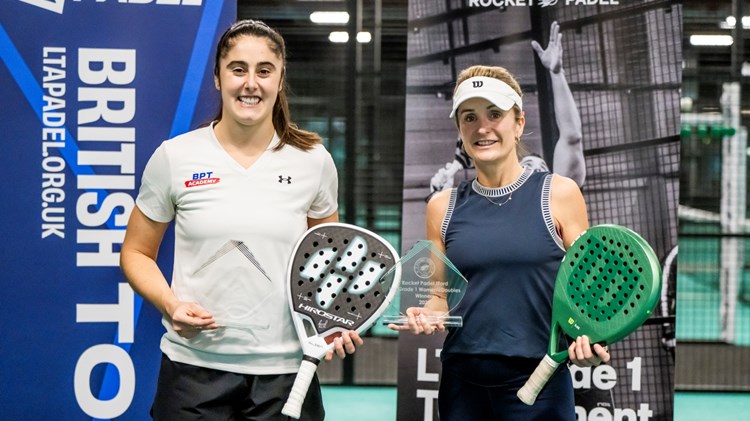 Two women holding padel bats and glass trophies wearing sports kit smile at the camera