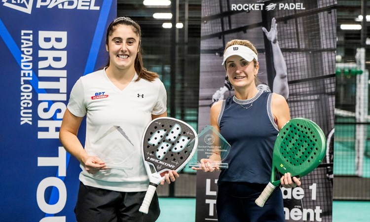 Two women holding padel bats and glass trophies wearing sports kit smile at the camera