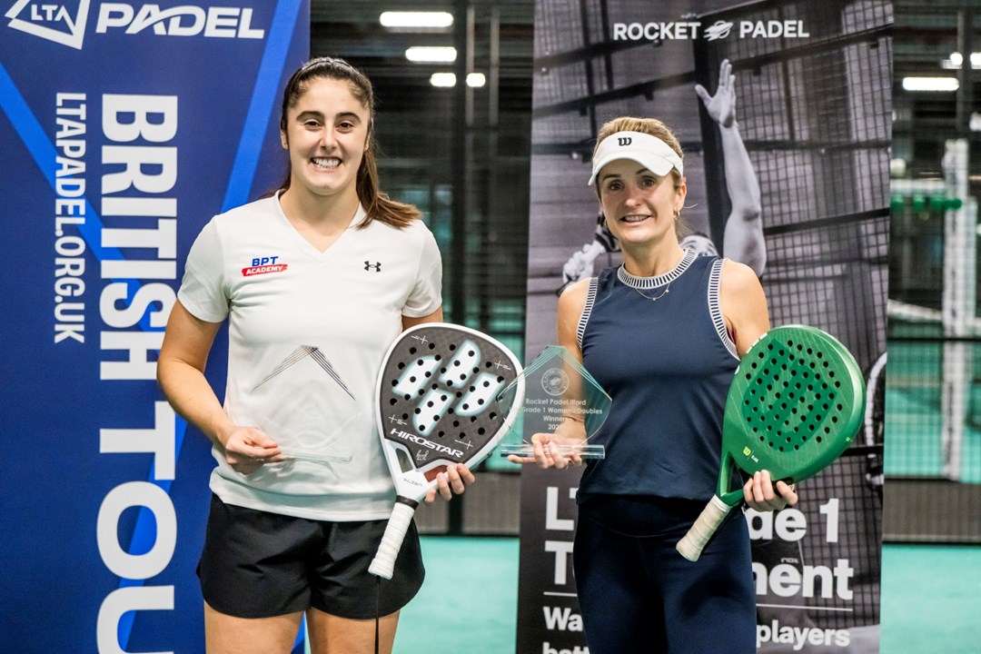 Two women holding padel bats and glass trophies wearing sports kit smile at the camera