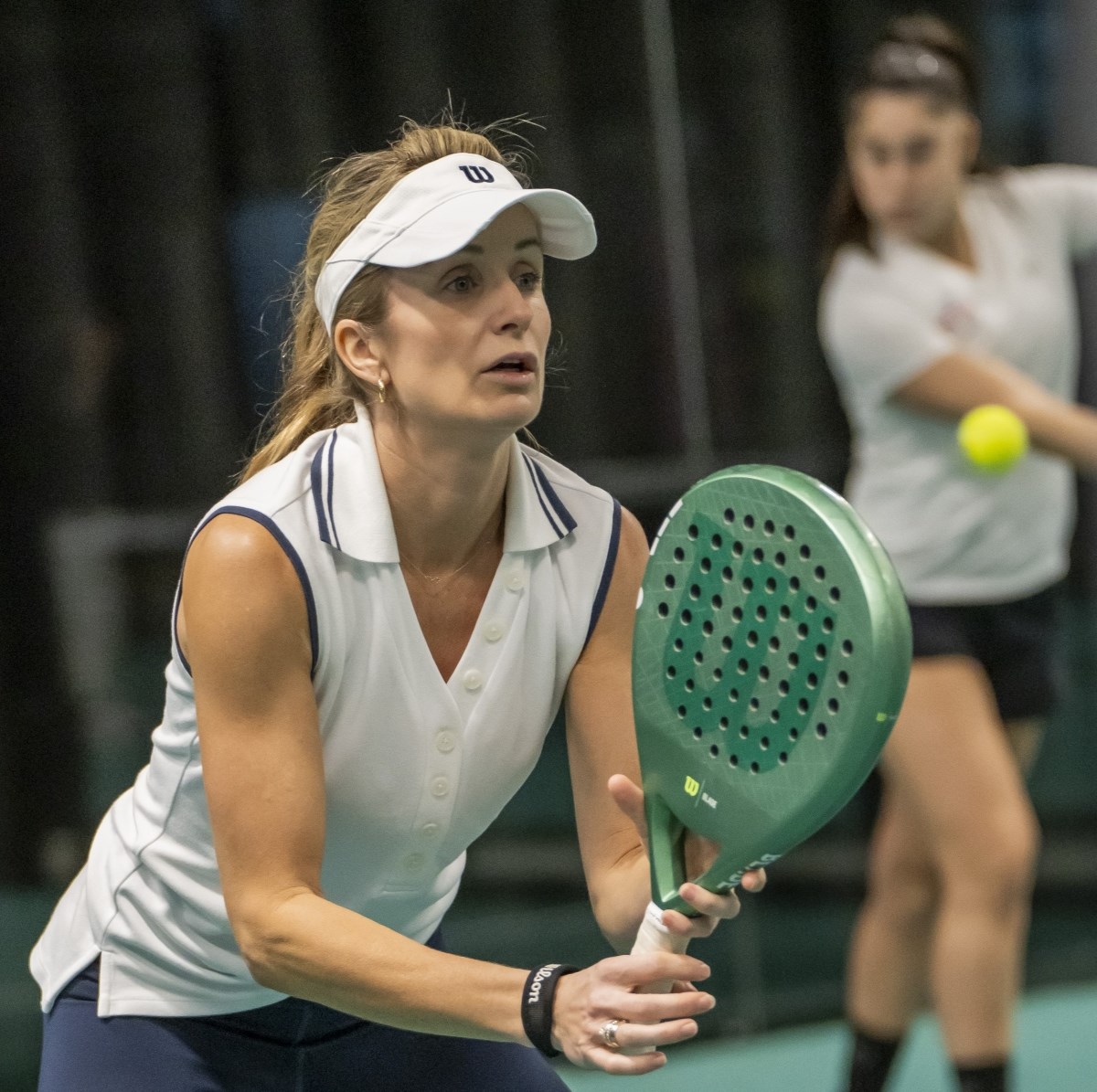 A woman wearing a white t-shirt holds a padel bat, whilst in the background, a person hits a tennis ball