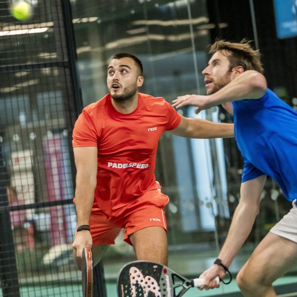 Two men, one wearing a red shirt and another wearing a blue shirt, run to hit a tennis ball in the air in front of them