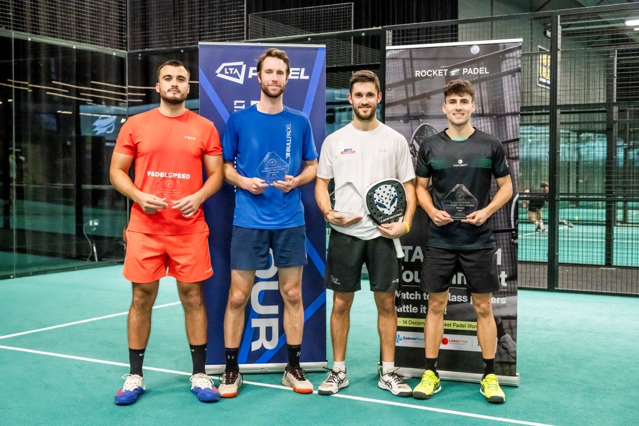 Four men sand holding glass trophies. Behidn them are two banners, with wire fencing behind those banners