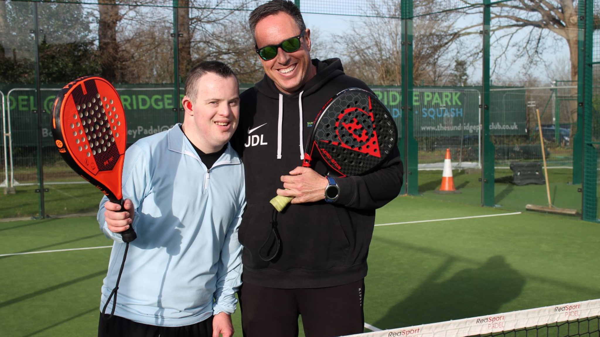 A young man with Downs Syndrome poses with a padel coach on an outdoor padel court