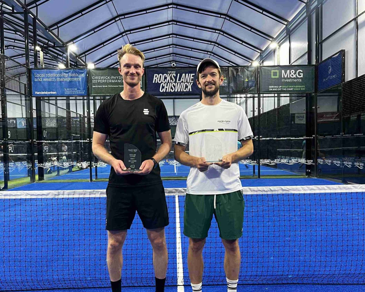 British padel players Nicholas Hatchett and Sam Macneil holding their trophies and smiling on court at the LTA Padel British Tour event at Rocks Lane, Chiswick