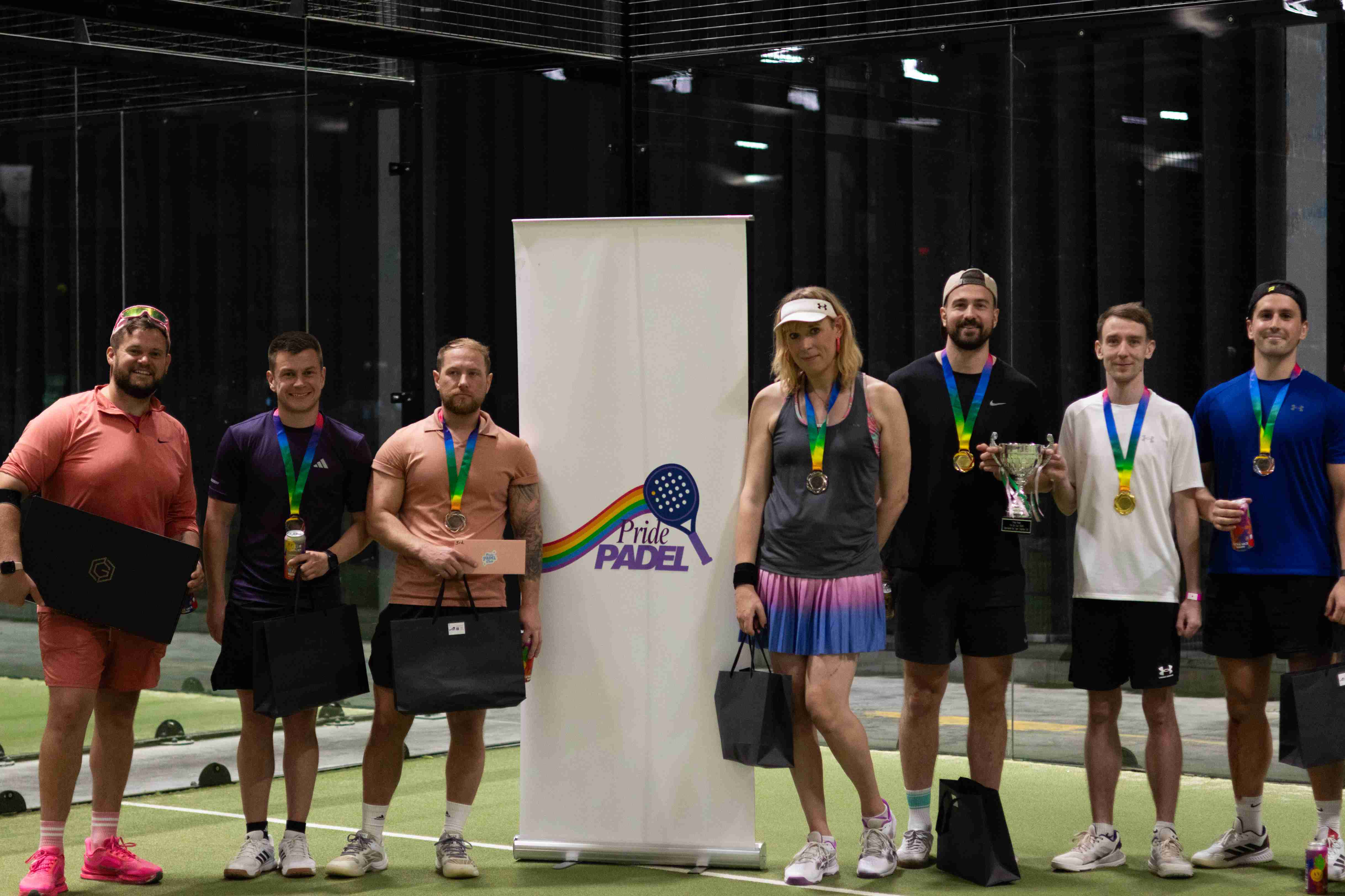 A group of male and female padel players stood in front of a sign saying 'Pride Padel'