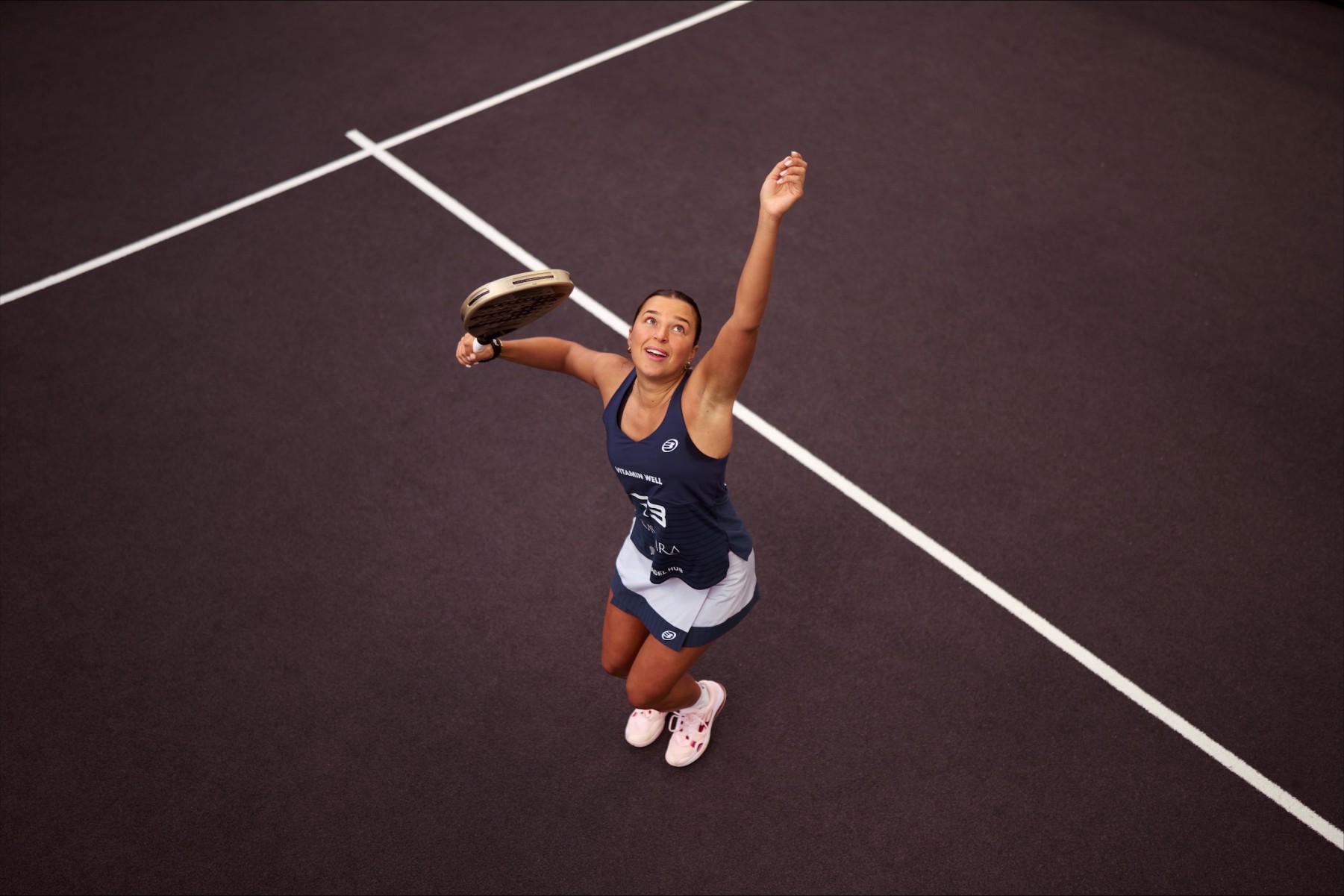 An ariel view shot of Catherine rose throwing a ball in the air and preparing to hit it with her padel racket on a padel court