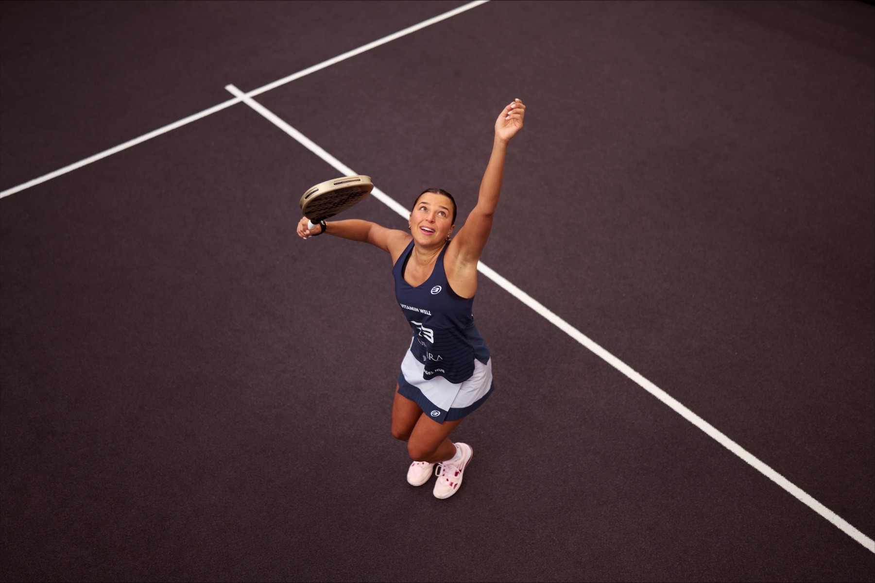 An ariel view shot of Catherine rose throwing a ball in the air and preparing to hit it with her padel racket on a padel court