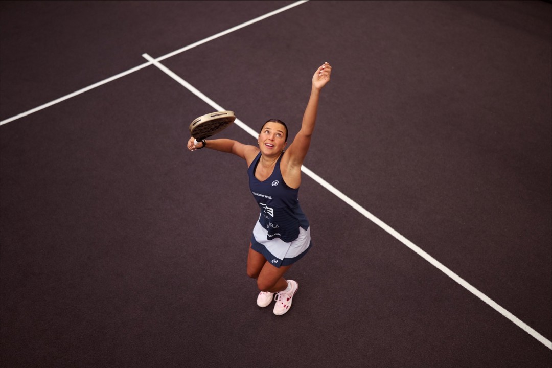 An ariel view shot of Catherine rose throwing a ball in the air and preparing to hit it with her padel racket on a padel court